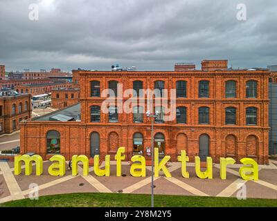 Bâtiment industriel en brique rouge rénové à Manufaktura, Łódź, Pologne, avec une signalisation jaune vif et une place urbaine un jour de printemps nuageux Banque D'Images