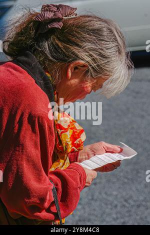 Une femme âgée dans une veste rouge et une écharpe à motifs lit une glissade omikuji à Hiroshima, au Japon. Ses cheveux grisonnés sont attachés. Un Struc flou Banque D'Images