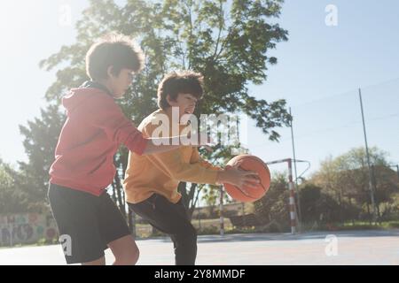 Deux garçons jouent au basket-ball sur un terrain extérieur, l'un tenant le ballon tandis que l'autre l'attrape. Ils sont habillés dans des sweats à capuche décontractés, Banque D'Images
