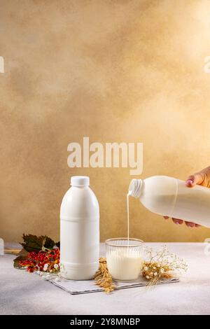 Verser à la main du lait ou du kéfir d'une bouteille dans un verre sur une table avec du blé et des fleurs Banque D'Images