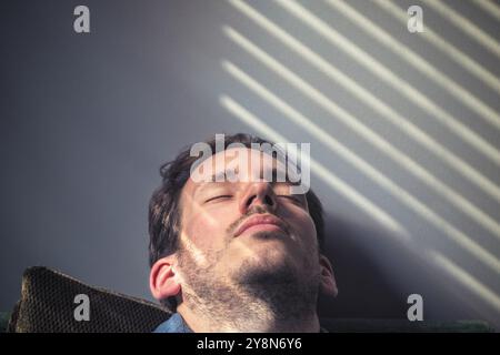 Portrait d'un homme caucasien somnolant sur le canapé, sur le visage tombe l'ombre des stores Banque D'Images