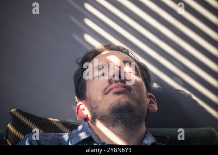 Portrait d'un homme caucasien somnolant sur le canapé, sur le visage tombe l'ombre des stores Banque D'Images