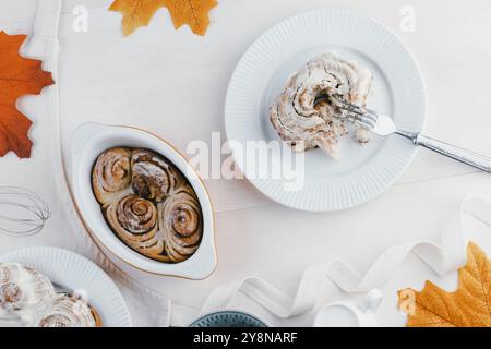Des petits pains à la cannelle fraîchement cuits reposent dans un plat à côté de pommes tranchées, d'un verre de lait et d'un bol de crème glacée, entourés de feuilles d'automne. Banque D'Images