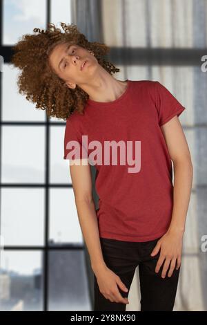 Cheveux secs et mats. Jeune homme en t-shirt rouge foncé luttant contre les cheveux bouclés emmêlés, debout dans le studio, espace de copie. Homme agitant sa coiffure bouclée. Banque D'Images