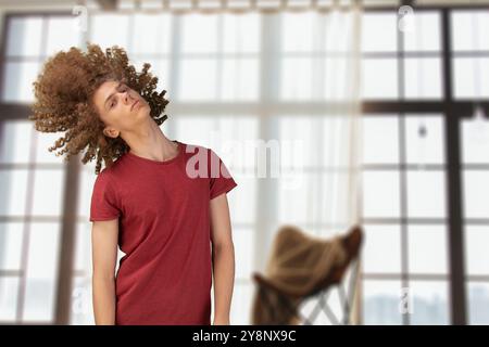 Homme agitant sa coiffure bouclée. Jeune homme en t-shirt rouge foncé luttant contre les cheveux bouclés emmêlés, debout dans le studio, espace de copie. Cheveux secs et mats. Banque D'Images