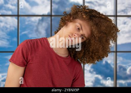 Jeune homme en t-shirt rouge foncé luttant contre les cheveux bouclés emmêlés, debout dans le studio, espace de copie. Homme agitant sa coiffure bouclée. Cheveux secs et mats. Banque D'Images