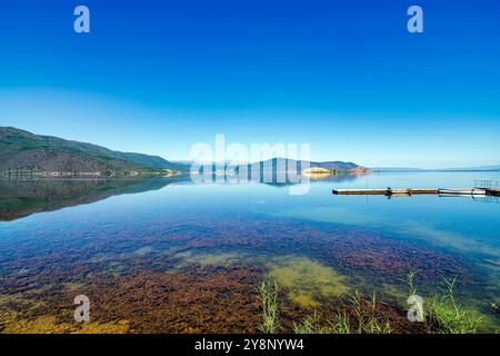 Eaux calmes du lac de Prespa au village de Mikrolimni, reflétant les montagnes environnantes avec de petits quais et des bateaux en vue. Banque D'Images