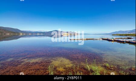 Eaux calmes du lac de Prespa au village de Mikrolimni, reflétant les montagnes environnantes avec de petits quais et des bateaux en vue. Banque D'Images