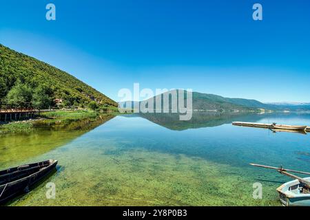 Eaux calmes du lac de Prespa au village de Mikrolimni, reflétant les montagnes environnantes avec de petits quais et des bateaux en vue. Banque D'Images