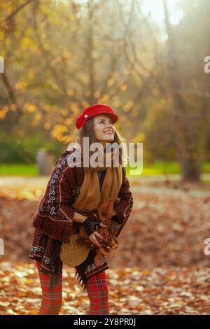 Bonjour l'automne. heureuse femme tendance de 40 ans en chapeau rouge avec écharpe, gants et sac jetant des feuilles d'automne dans le parc de la ville. Banque D'Images