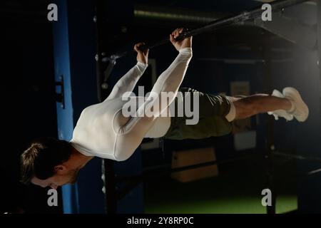 Sportif en forme effectuant un exercice de levier arrière avec poignée prononcée maintenant le corps droit de tension des muscles tout en s'entraînant sur la barre haute dans une salle de gym faiblement éclairée, espace de copie Banque D'Images