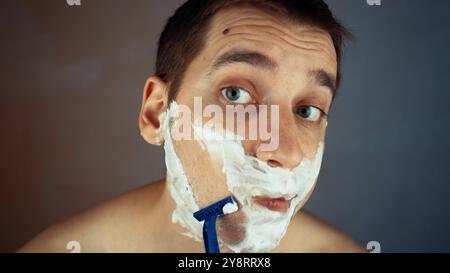 Un gars d'apparence européenne, un bel homme barbu se rase avec un rasoir jetable dans la salle de bain, regardant dans le miroir. Routine quotidienne du matin masculin Banque D'Images