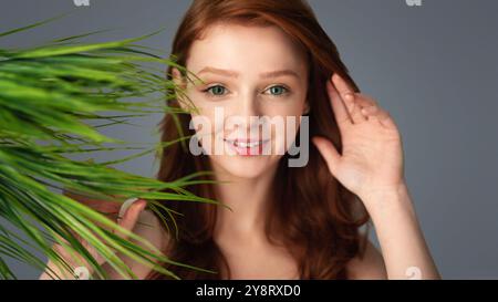 Jeune femme aux cheveux de gingembre posant avec des feuilles de plantes vertes, fond gris Banque D'Images