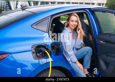 Jeune femme blonde parle sur son smartphone pendant que sa voiture électrique se recharge à une station de VE Banque D'Images