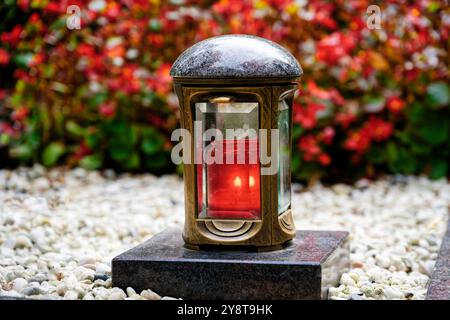 une lanterne funéraire en laiton dans un cimetière avec une bougie rouge allumée sur un piédestal en marbre au milieu de galets blancs devant des plantes rouges dans un fond flou Banque D'Images