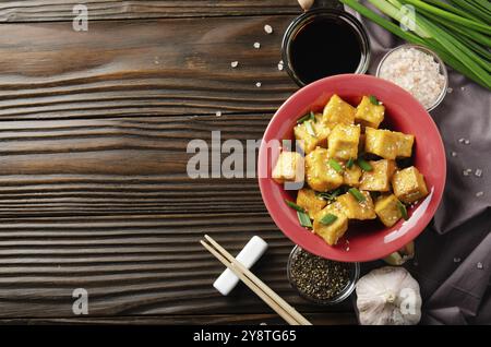 Vue sur la couche plate avec des cubes de tofu sautés avec des ciboulettes dans un plat d'argile sur une table de cuisine en bois avec des ciboulettes de serviette à l'ail au poivre et sauce soja sur place Banque D'Images