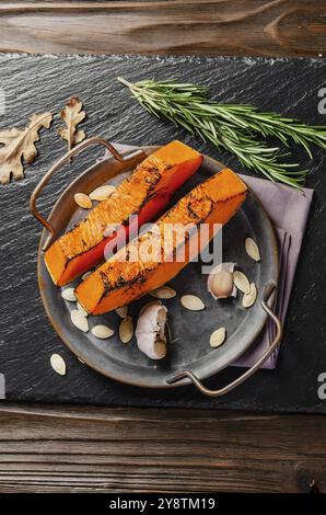 Vue à plat sur les tranches de citrouille cuites sur un plateau en métal sur une table en bois rustique de cuisine avec gousses d'ail et romarin Banque D'Images