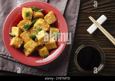 Vue sur la couche plate avec des cubes de tofu frits et croustillants, sautés et sautés dans un plat d'argile sur une table de cuisine en bois avec serviette et sauce soja sur place Banque D'Images