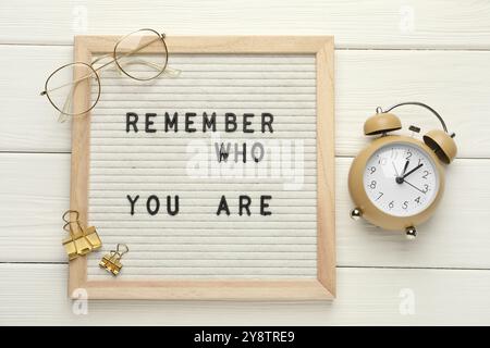 Tableau aux lettres avec phrase rappelez-vous qui vous êtes, réveil, clips et verres sur table en bois blanc, pose à plat Banque D'Images