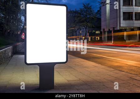 Maquette blanche vierge de panneau d'affichage vertical d'affiche avec des sentiers de lumière de voiture la nuit. Publicité vide bannière extérieure sur City Street Banque D'Images