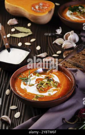 Soupe de citrouille rustique maison avec des graines dans un plat d'argile sur une table en bois avec des épices à pain et des greens de côté Banque D'Images