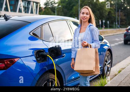 Femme se tient à côté de sa voiture électrique, la chargeant et tenant un sac en papier de provisions Banque D'Images