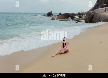 Une femme est assise sur une plage de sable avec l'océan et de grands rochers en arrière-plan. Banque D'Images