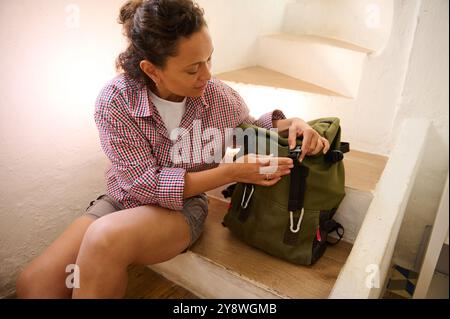 Une femme assise dans les escaliers, organisant un sac à dos vert, préparant un voyage ou une aventure en plein air. Ambiance décontractée et concentrée. Banque D'Images