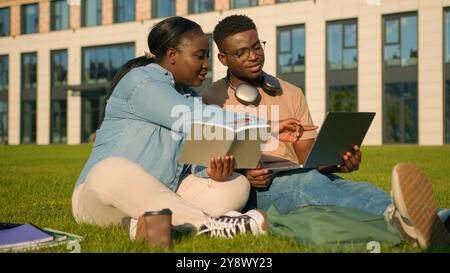 Afro-américaine jeunes étudiantes filles amis les élèves sont assis à l'herbe verte dans le parc étudiant avec ordinateur portable e-learning discuter Banque D'Images