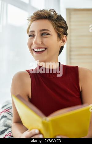 Une jeune femme aux cheveux courts aime lire un livre à la maison, rayonnant de joie dans sa routine quotidienne. Banque D'Images