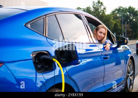 Femme blonde se penche par la fenêtre pendant que le véhicule électrique se recharge à une station dans la rue Banque D'Images