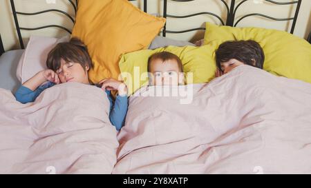 Trois enfants allongés sur un lit. Deux dorment et le plus jeune est éveillé. Le bébé regarde la caméra Banque D'Images