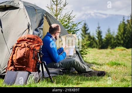 L'homme est assis dans l'entrée de la tente touristique, étudiant la carte avec une expression réfléchie. À côté de lui sac à dos orange, équipé d'un trépied et d'un panneau solaire, placé sur fond pittoresque d'arbres et de montagnes. Banque D'Images