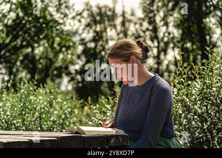 Femme assise tranquillement à une table en bois, engrossée dans un livre tout en étant entourée de verdure dans un cadre extérieur, profitant de la lecture dans un moment serein Banque D'Images