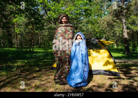 Famille de touristes d'un père et d'une fille posent et dansent drôle dans des sacs de couchage près d'une tente. Loisirs de plein air en famille, camping, équipement de randonnée Banque D'Images