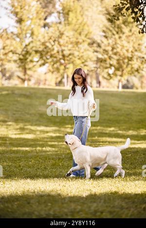 Une jeune femme en tenue d'automne promène joyeusement son chien dans un parc animé rempli d'arbres. Banque D'Images