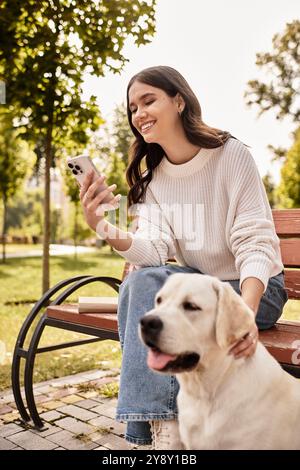 Assise sur un banc dans le parc, une jeune femme sourit en caressant son chien et en utilisant son téléphone. Banque D'Images