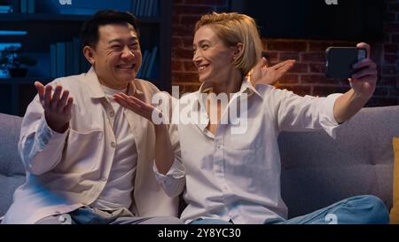 Heureux excité multiracial couple multiethnique marié à divers hommes asiatiques et femme caucasienne parlant en ligne conférence par appel vidéo avec application de téléphone mobile Banque D'Images