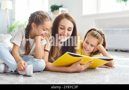 Famille heureuse allongée sur le sol à la maison, mère et filles souriantes appréciant la lecture du livre ensemble. Banque D'Images