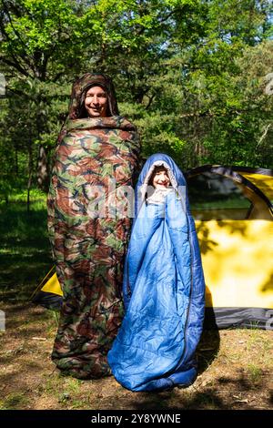 Famille de touristes d'un père et d'une fille posent et dansent drôle dans des sacs de couchage près d'une tente. Loisirs de plein air en famille, camping, équipement de randonnée Banque D'Images