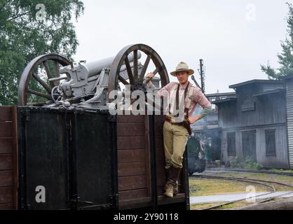 Un jeune homme vêtu d'une tenue historique pose en toute confiance au sommet d'un wagon vintage arborant un canon. La scène capture une journée nuageuse à un historica Banque D'Images