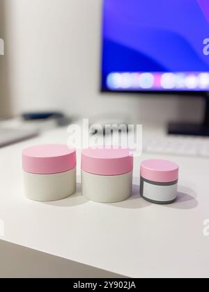 Configuration de bureau minimaliste avec trois pots de soins de la peau avec couvercles roses, parfaitement alignés sur un fond de technologie moderne. Copier l'espace. Élégant Banque D'Images