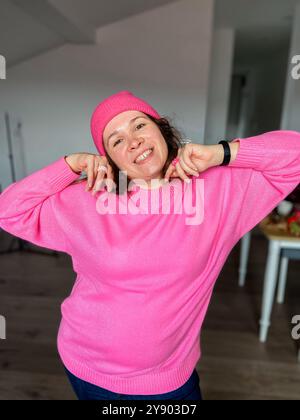 Femme millénaire pose joyeusement dans un pull rose vif et chapeau assorti, exsudant le bonheur et la confiance dans un cadre décontracté à la maison. Joyeux Banque D'Images