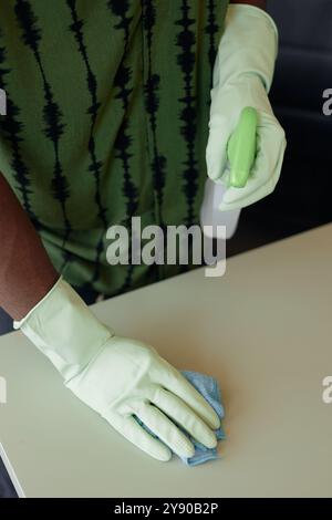 Vue de dessus minimale gros plan d'un homme afro-américain méconnaissable nettoyant la table et portant des gants tout en pulvérisant du liquide désinfectant, espace de copie Banque D'Images
