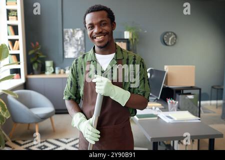 Portrait de taille haute de jeune homme afro-américain souriant nettoyant le bureau et regardant l'appareil photo tenant la vadrouille, l'espace de copie Banque D'Images