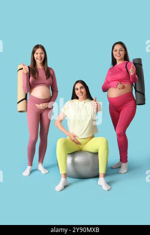 Belles jeunes femmes enceintes sportives avec ballon de fitness et tapis de yoga montrant un geste pouce vers le haut sur fond bleu Banque D'Images