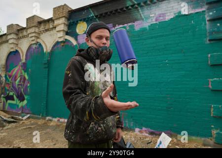 Guy dans respirateur, bonnet et sweat à capuche jetant la boîte bleue avec de la peinture tout en se tenant contre le mur vert du bâtiment Banque D'Images