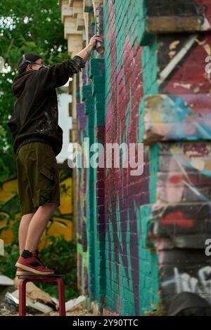 Vue latérale d'un jeune homme sous respirateur debout sur un tabouret devant le mur du bâtiment et pulvérisant de la peinture à partir du distributeur Banque D'Images