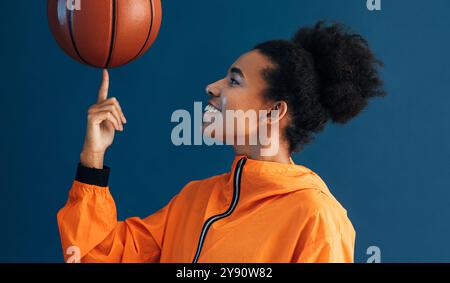 Vue de côté d'une femme souriante filant du basket-ball sur son doigt tout en posant en studio sur fond bleu Banque D'Images