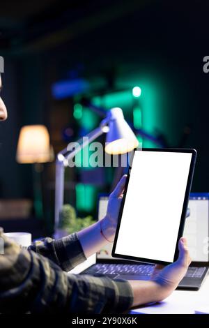 Personne tenant verticalement la tablette avec écran blanc devant l'ordinateur portable. Freelance masculine travaillant à la maison, axée sur l'appareil numérique affichant un modèle de maquette chromakey vierge. Banque D'Images